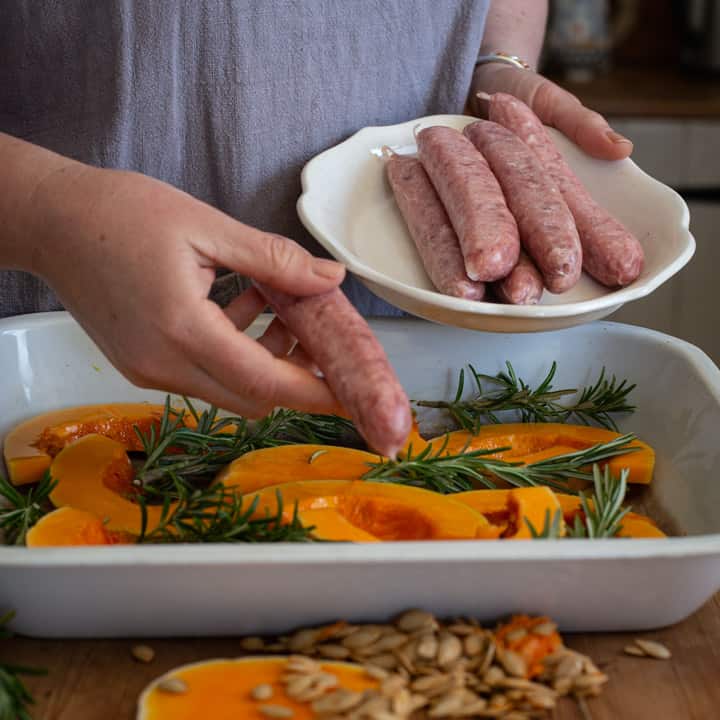 womans hands placing butchers sausages on slices of veggies in a white enamel baking pan