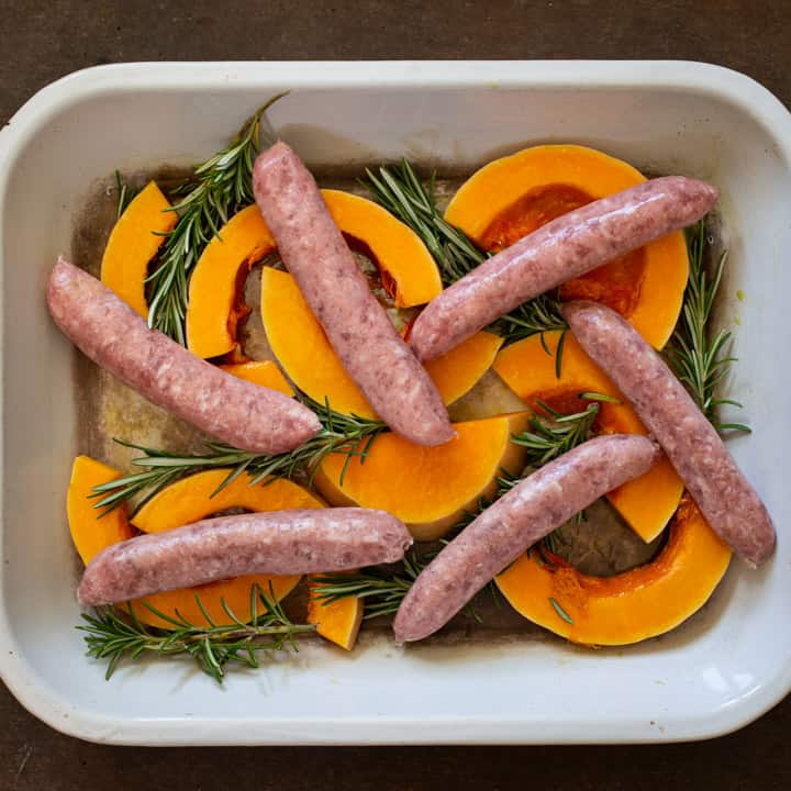 overhead shot of white enamel baking tin with orange squash slices, green sprigs of rosemary and 6 sausages