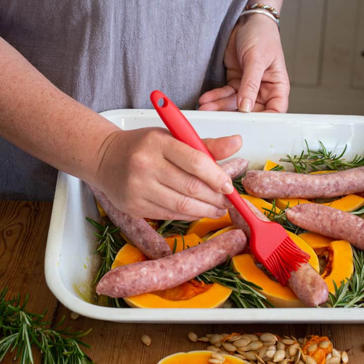 womans hands brushing a tray of sausages and veggies with oil on a red silicone brush