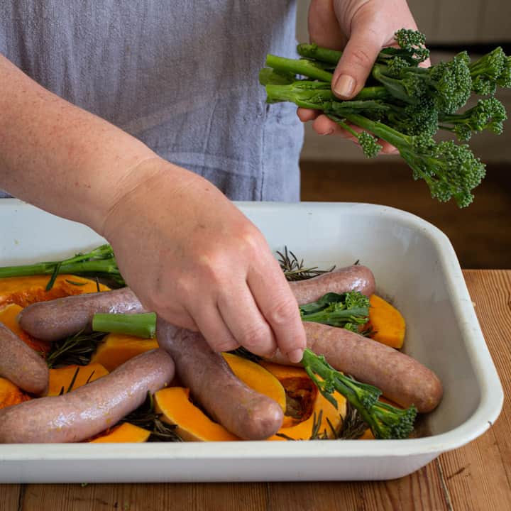 womans hands placing pieces of tenderstem broccoli onto a white tray of sausages and veggies