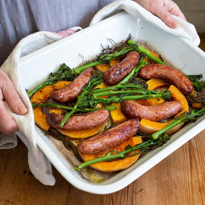 woman in grey holding a white enamel sheet pan with sausages, broccolini and butternut squash