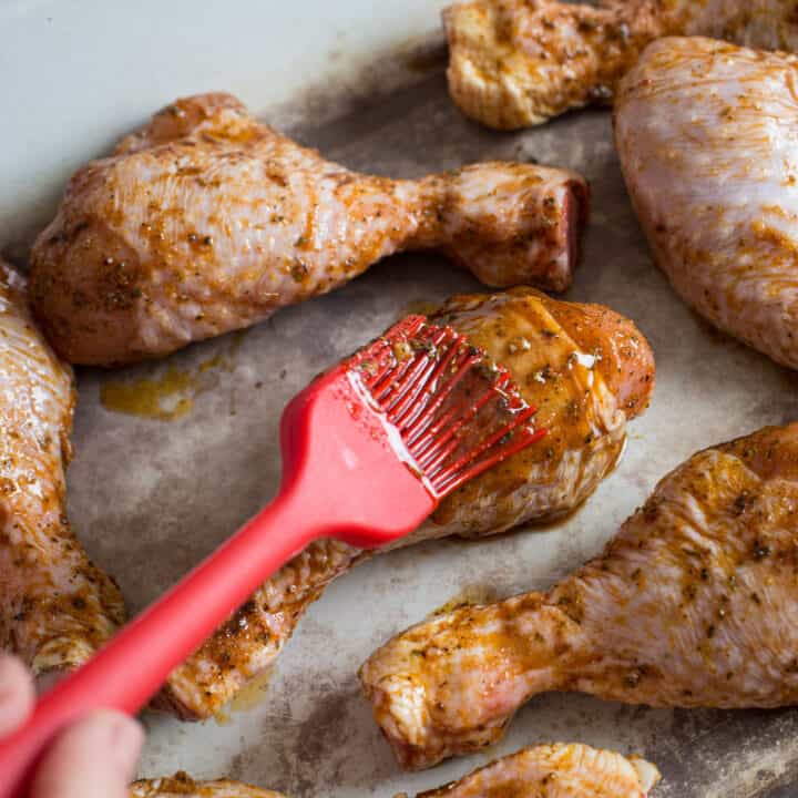 White enamel baking tray with several marinated chicken drumsticks and a red silicone brush adding more marinade