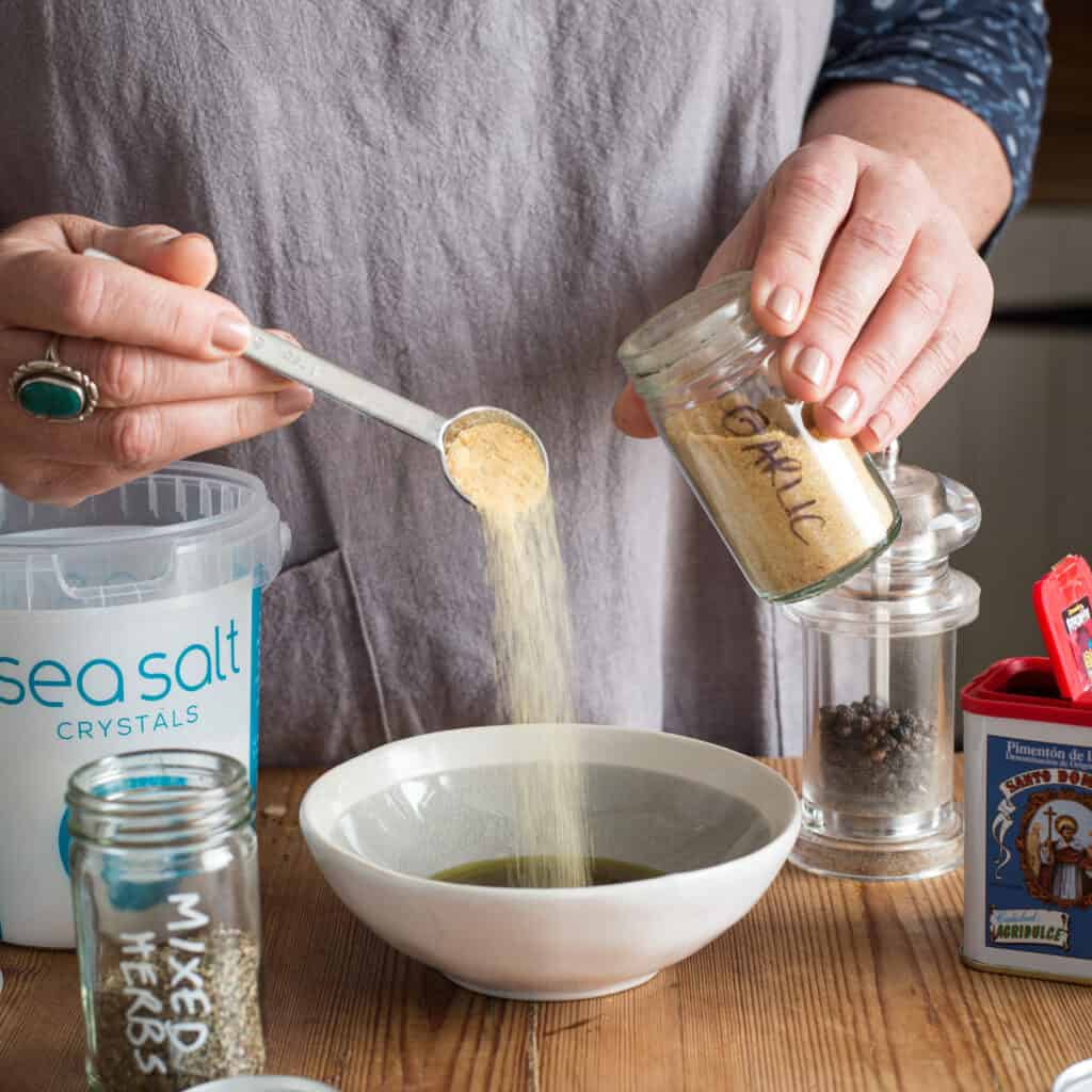 Woman in grey tipping a teaspoon of garlic into a small bowl