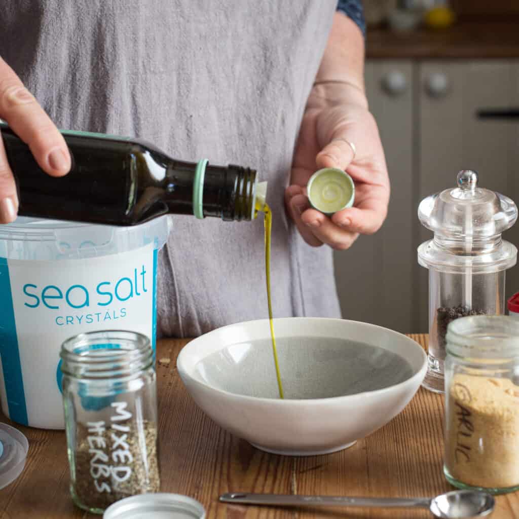 Woman pouring oil from a dark bottle into a smal white and grey bowl on a wooden kitchen counter