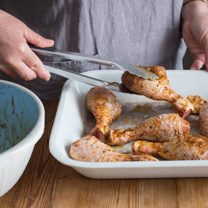 Woman in grey placing marinated chicken drumsticks onto a white enamel baking tray 