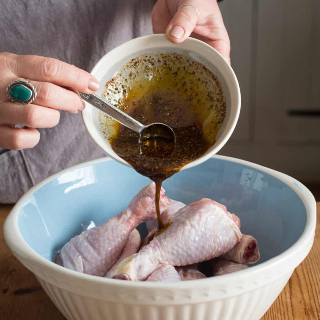Woman’s hands pouring marinade from a small grey bowl into a large bowl of chicken drumsticks