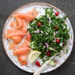 White plate with bright green broccoli kale salad, lemon slice and smoked salmon slices