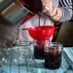 Woman pouring hot blackberry jam into a red plastic jam funnel over a glass jar