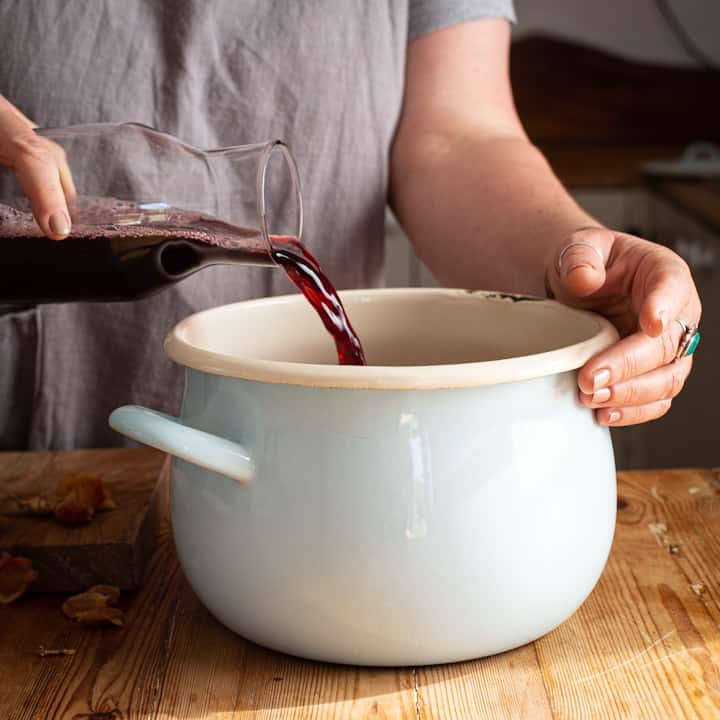 Woman in grey pouring red grape juice from a glass bottle into a blue and white saucepan