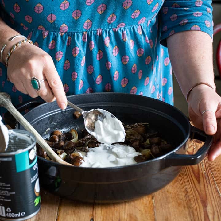 Woman in blue stirring coconut milk into a black pan of mushrooms