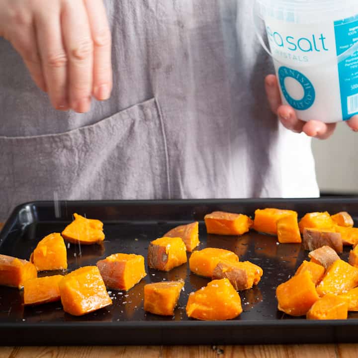Woman in grey sprinkled diced sweet potatoes on a black baking try with Cornish sea salt