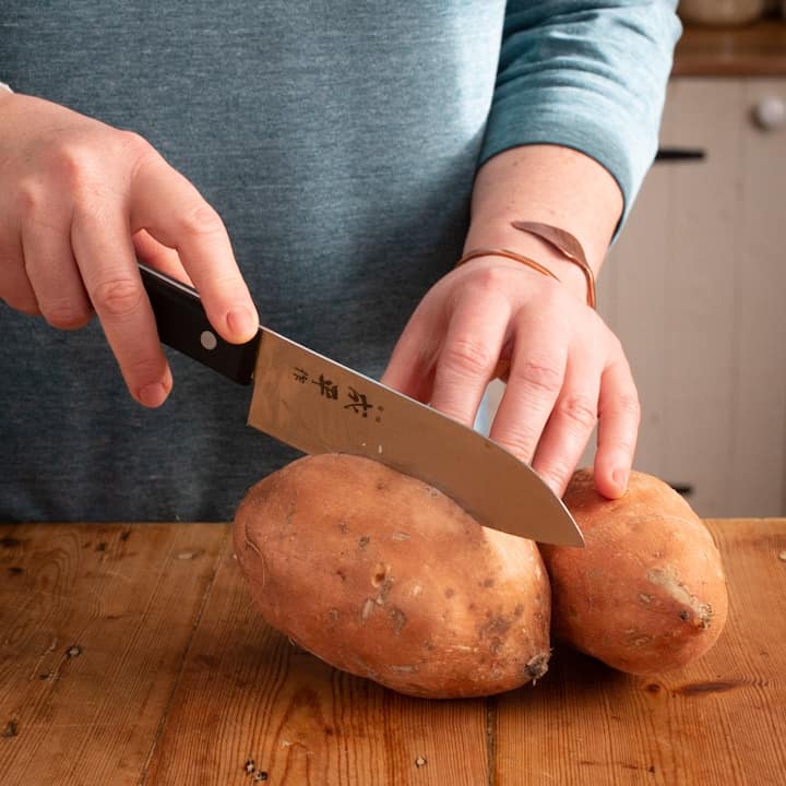 Woman hands using a black handled Japanese knife to score 2 large sweet potatoes before baking