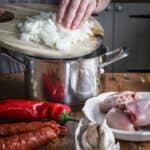 Woman’s hands pouring chopped onions from a wooden chopping board into a silver saucepan surrounded by chicken stew ingredients