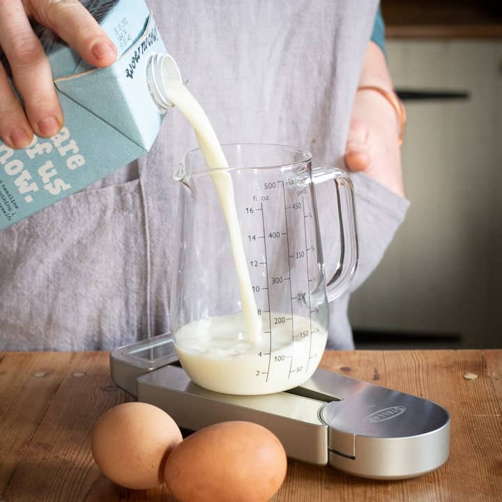 Woman’s hands pouring oat milk from a pale blue carton into a glass jug on a silver set of kitchen scales