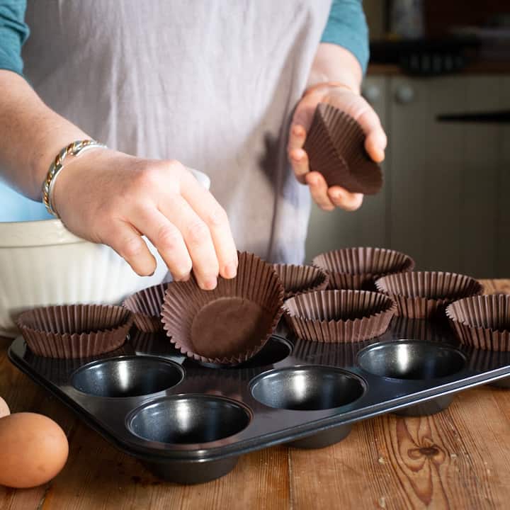 Woman’s hands placing brown paper muffin cases into a black muffin pan on a wooden counter