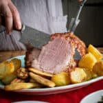 Woman carving a ham joint on a red tablecloth with roast vegetables and sides