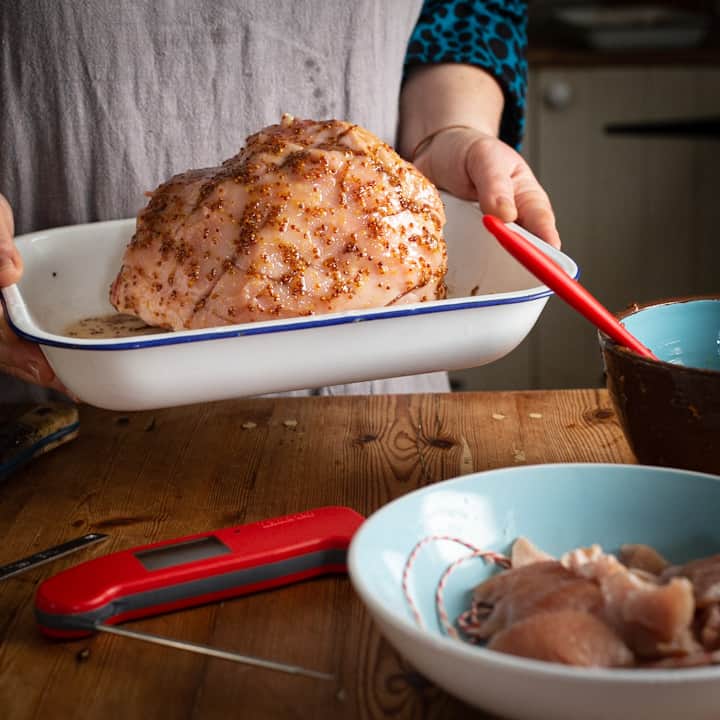 Woman in grey holding a white baking dish with a boiled ham coated in mustard and honey glaze ready for teh oven