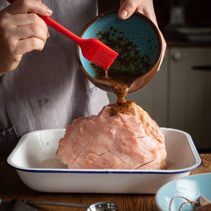 Woman in grey pouring a homemade glaze over a ham from a blue and brown bowl