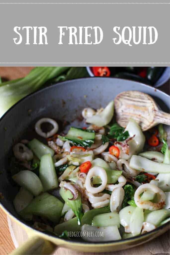 Inside shot of a grey frying pan showing a stir fry squid dish with pak Choi, red chilli peppers and a wooden spoon