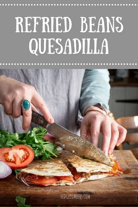 Womans hands slicing a refried beans quesadilla into triangle with a large silver knife surrounded by tomatoes and fresh coriander