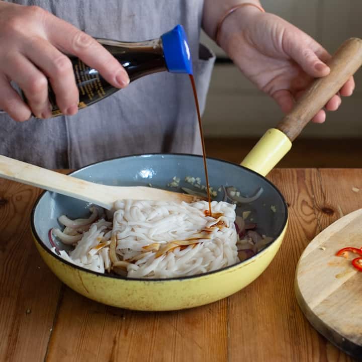 Womans hands pouring soy sauce into a yellow frying pan of noodles