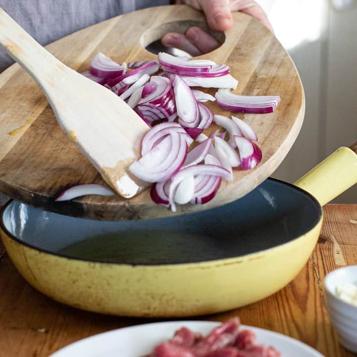 Red onions slices being scraped from a wooden chopping board into a yellow frying pan
