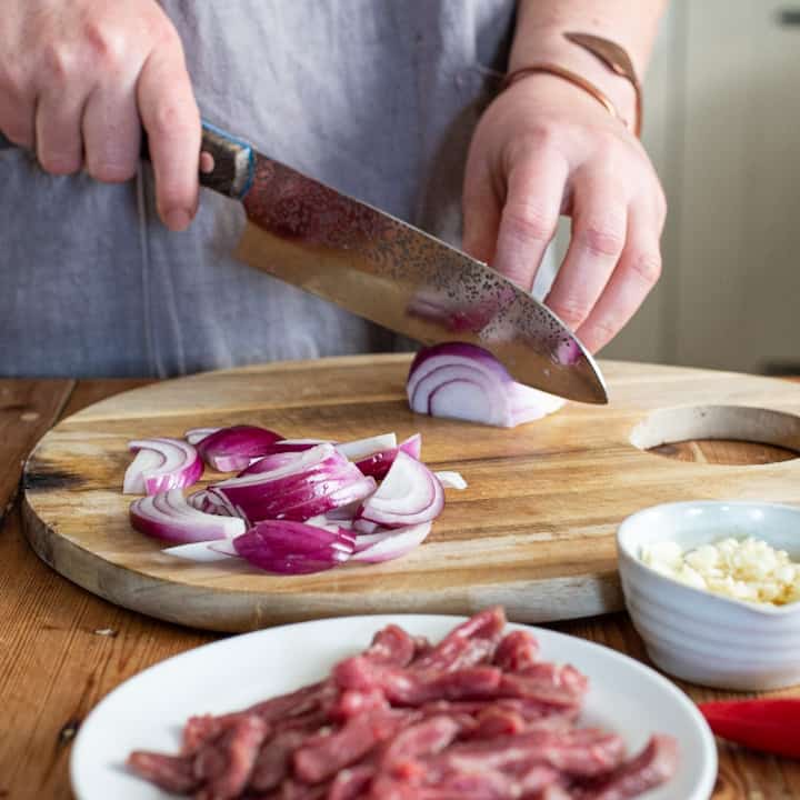 Womans hands slicing red onion on a wooden chopping board with a large knife