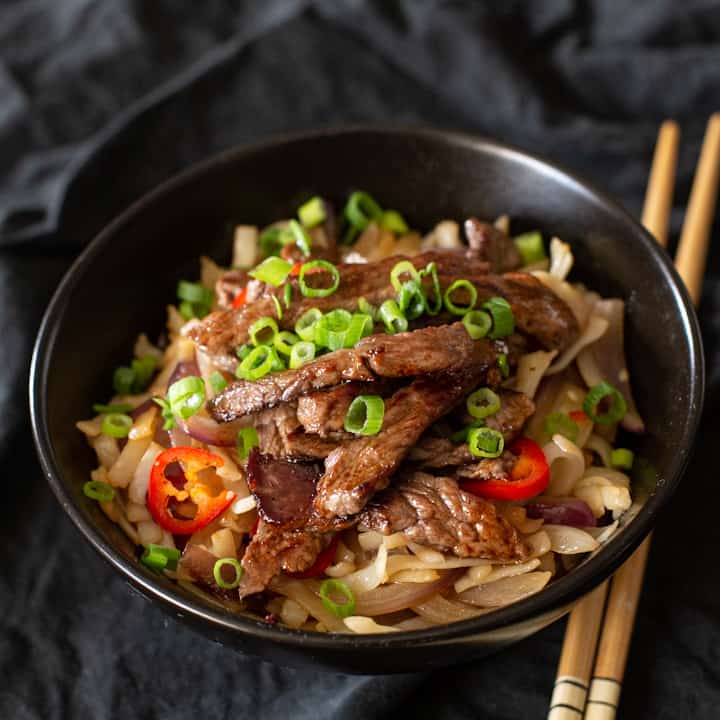 Black bowl with chopsticks and a portion of pan friend beef steak, noodles, red chilli and spring onions
