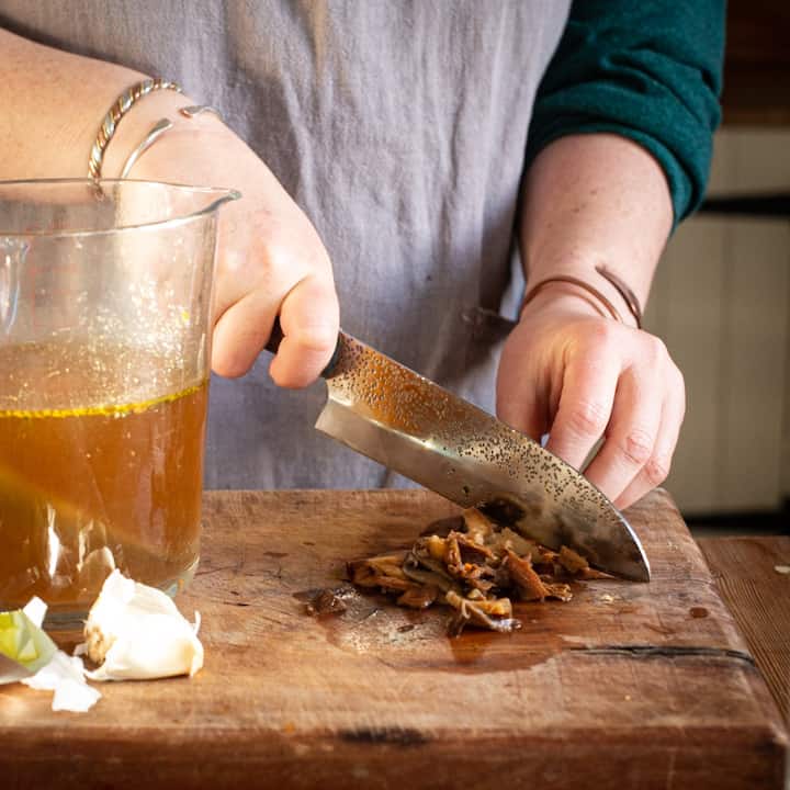 Woman’s hands chopping soaked mushrooms on a wooden chopping board next to a large glass just of mushroom broth