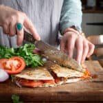 Womans hands slicing a refried beans quesadilla into triangle with a large silver knife surrounded by tomatoes and fresh coriander