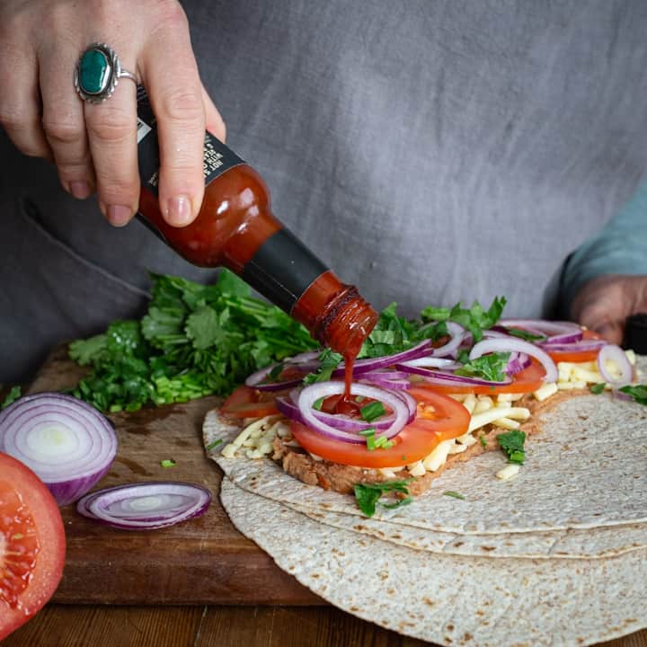 Womans hands pouring bright red hot sauce from a small bottle into a tortilla filled with refried beans and fresh veggies