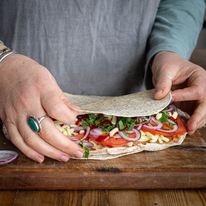 Woman’s hands folding a wheat tortilla over a mix of cheese, veggies and refried beans on a wooden board