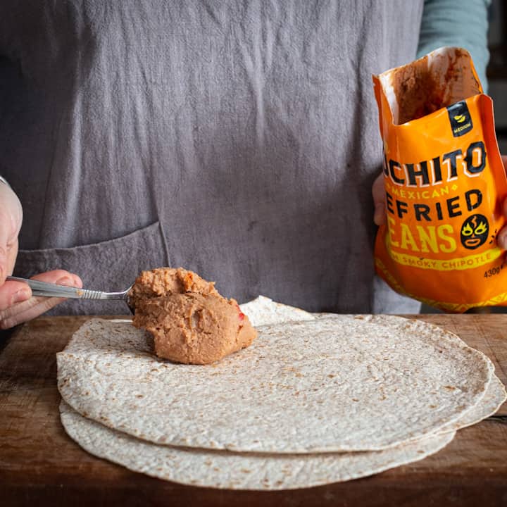 Womans hands spooning refried beans from an orange pouch with a silver spoon onto a wheat tortilla