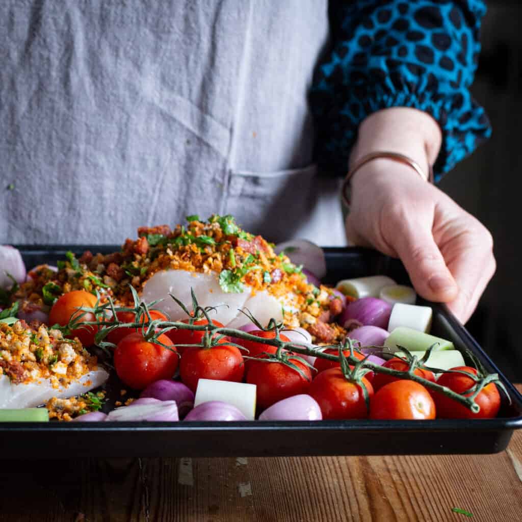 Woman in grey holding a black baking tray with fresh veggies and two pieces of cod topped with a spicy crumb