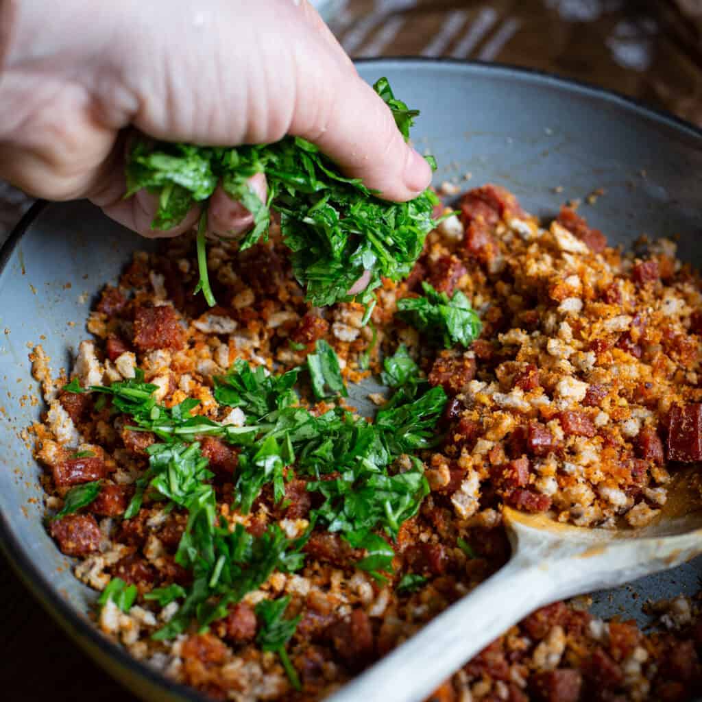 Hand sprinkling parsley into a frying pan of chorizo crumb topping