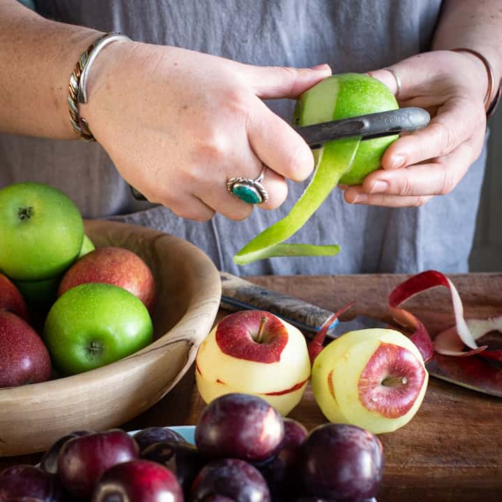 Woman’s hands peeling a green apple with a black peeler with several more apples and plums in the bottom of the image