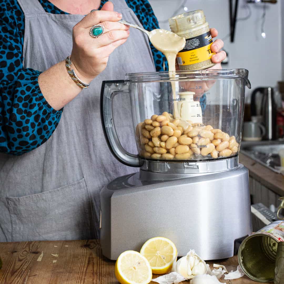 Woman in grey pinky pouring tahini from a silver spoon into a food mixer