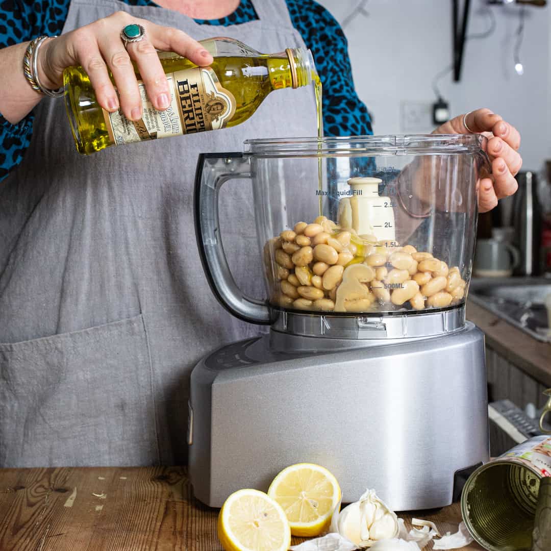 Woman pouring olive oil into a food processor filled with butter beans
