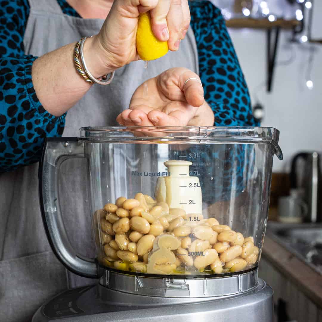 Woman in grey squeezing a lemon into a mixer of hummus ingredients