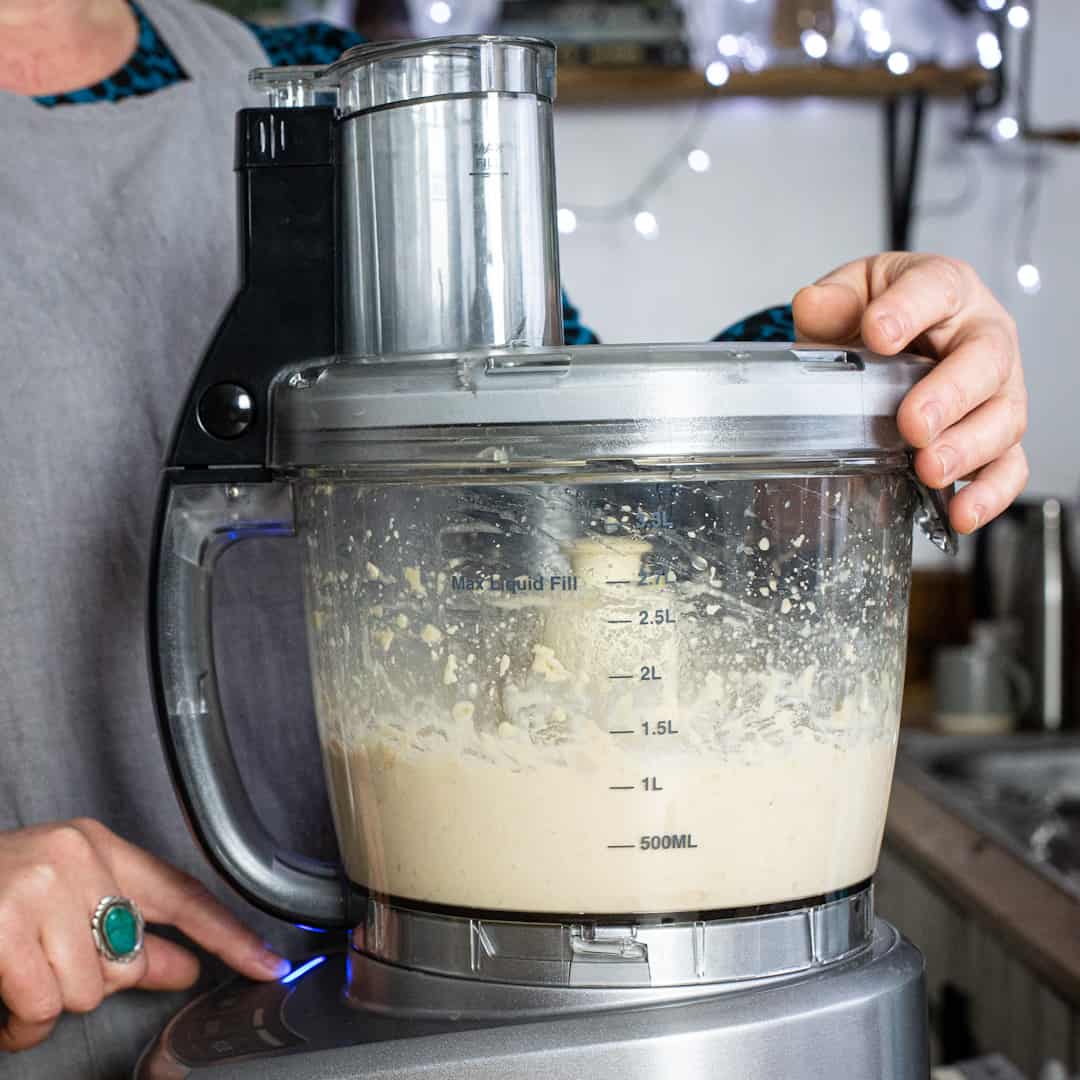 Woman in grey mixing cooked beans in a silver food processor to make hummus