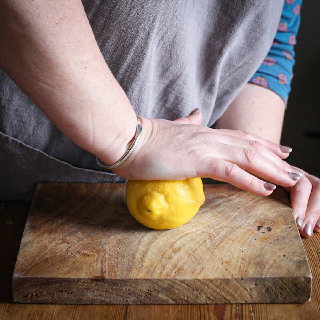 Woman in grey rolling a lemon with the heel of her hand on a wooden chopping board