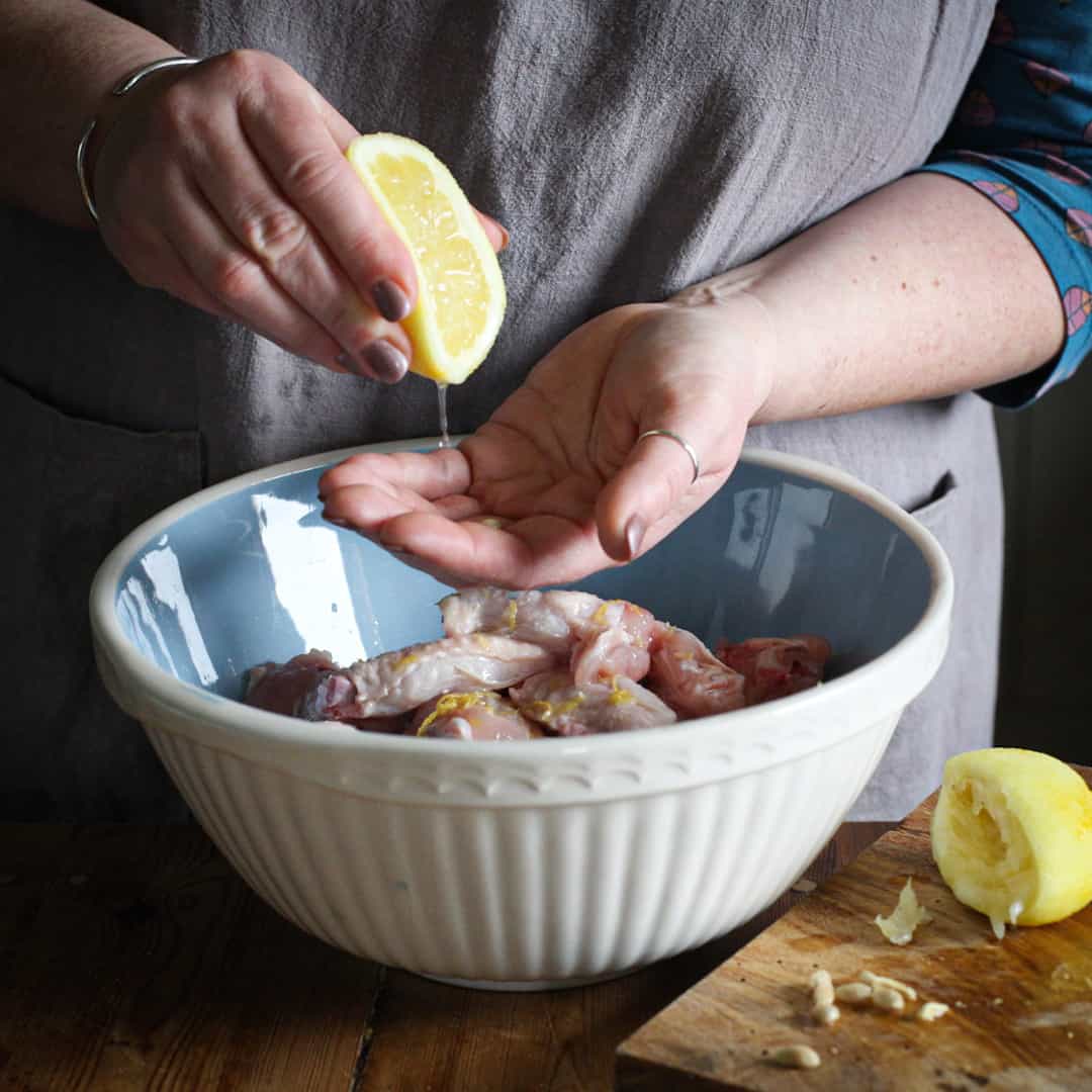 Woman in grey squeezing juice from half a lemon through her hands into a large white bowl of chicken wings