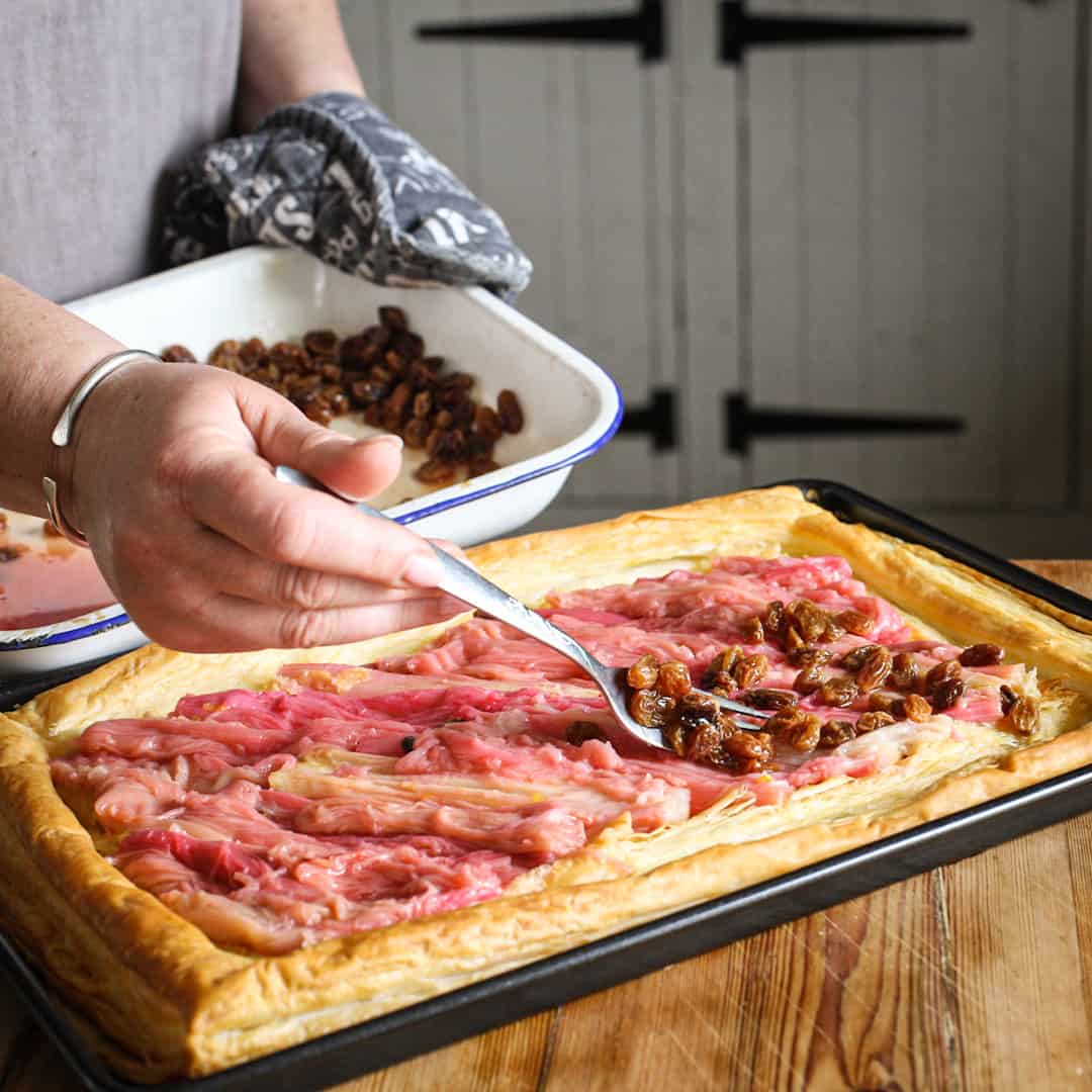 Womans hands spooning juicy sultanas onto baked rhubarb on a puff pastry base