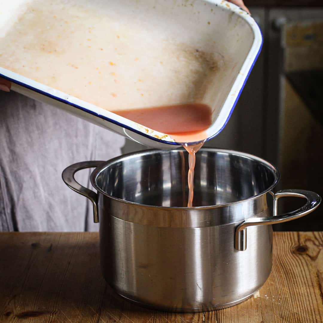 White enamel baking tray of fruit juices being poured into a silver saucepan on a rustic kitchen counter