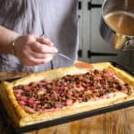 Woman spooning thickened fruit juices from a silver pan with a silver spoon over a puff pastry rhubarb tart