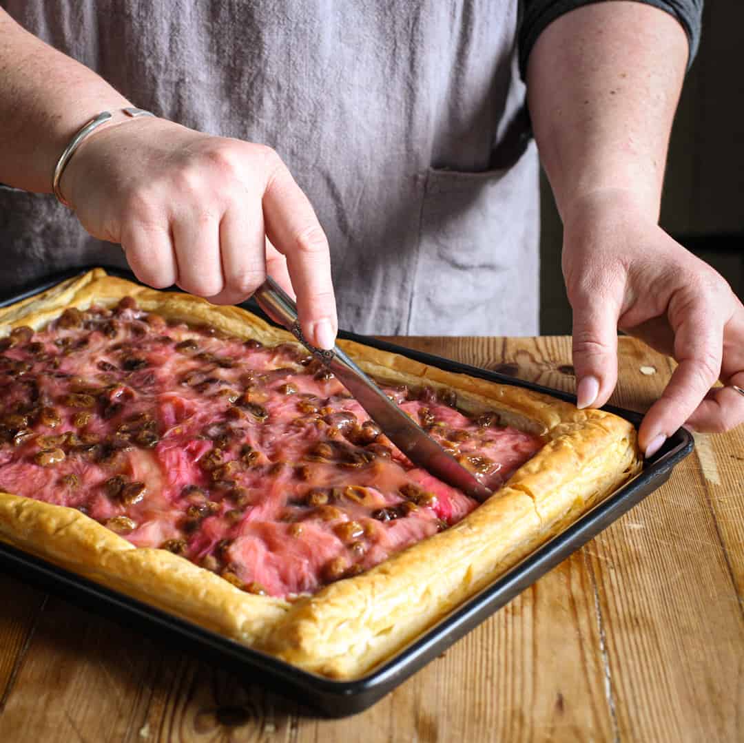 Womans hands slicing a hot, fresh baked rhubarb slice on a black baking tray