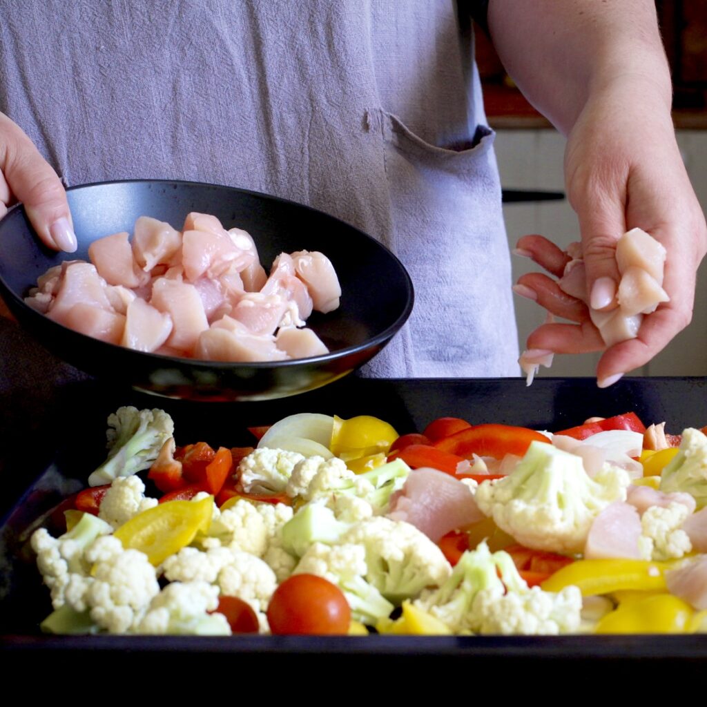 Woman holding a black bowl of chopped chicken in one hand, whilst scattering chicken pieces over a black baking tray of chopped veggies