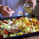 Woman in grey scattering a homemade spice blend over a baking tray of chopped veggies and chicken