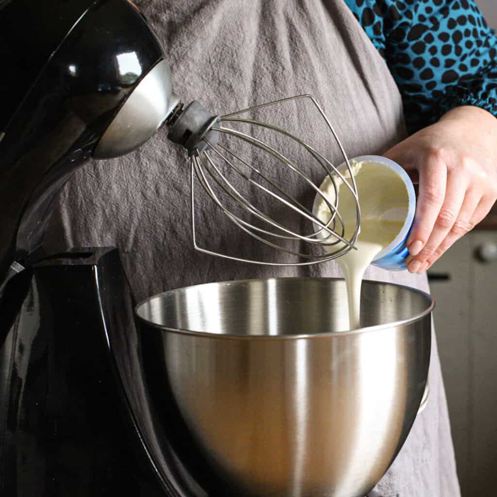 Woman pouring double cream into a large silver mixing bowl from plastic white and blue tub of cream