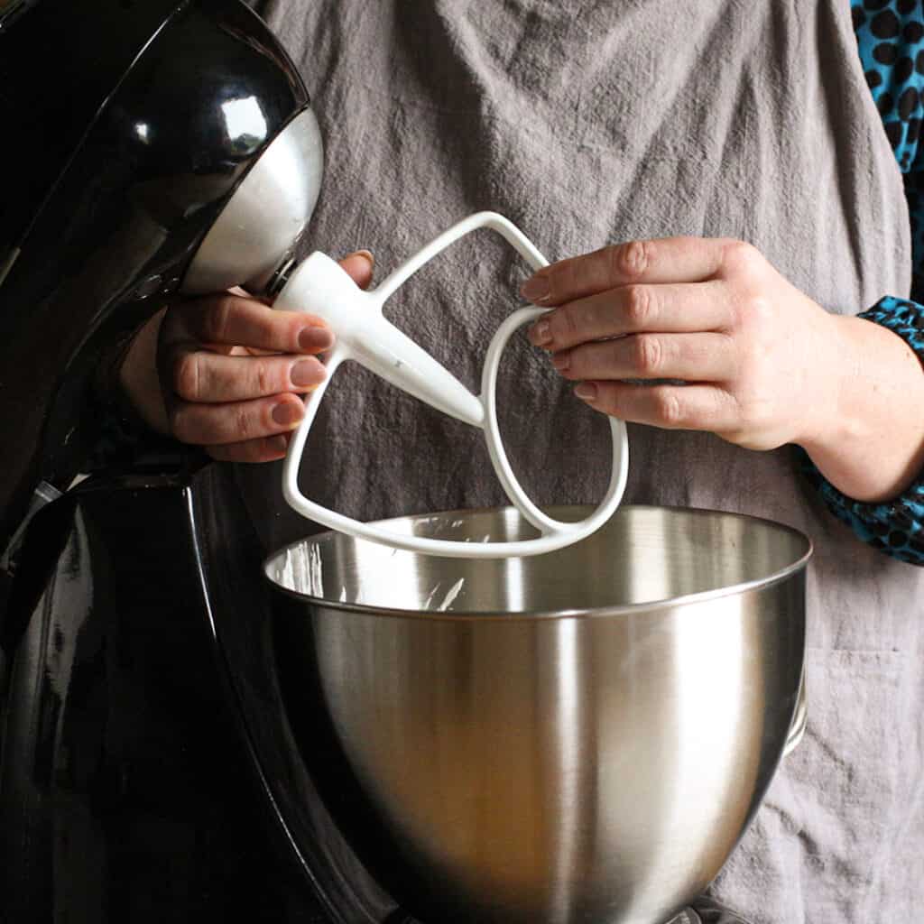 Woman in grey holding a dough hook over s stainless steel mixing bowl